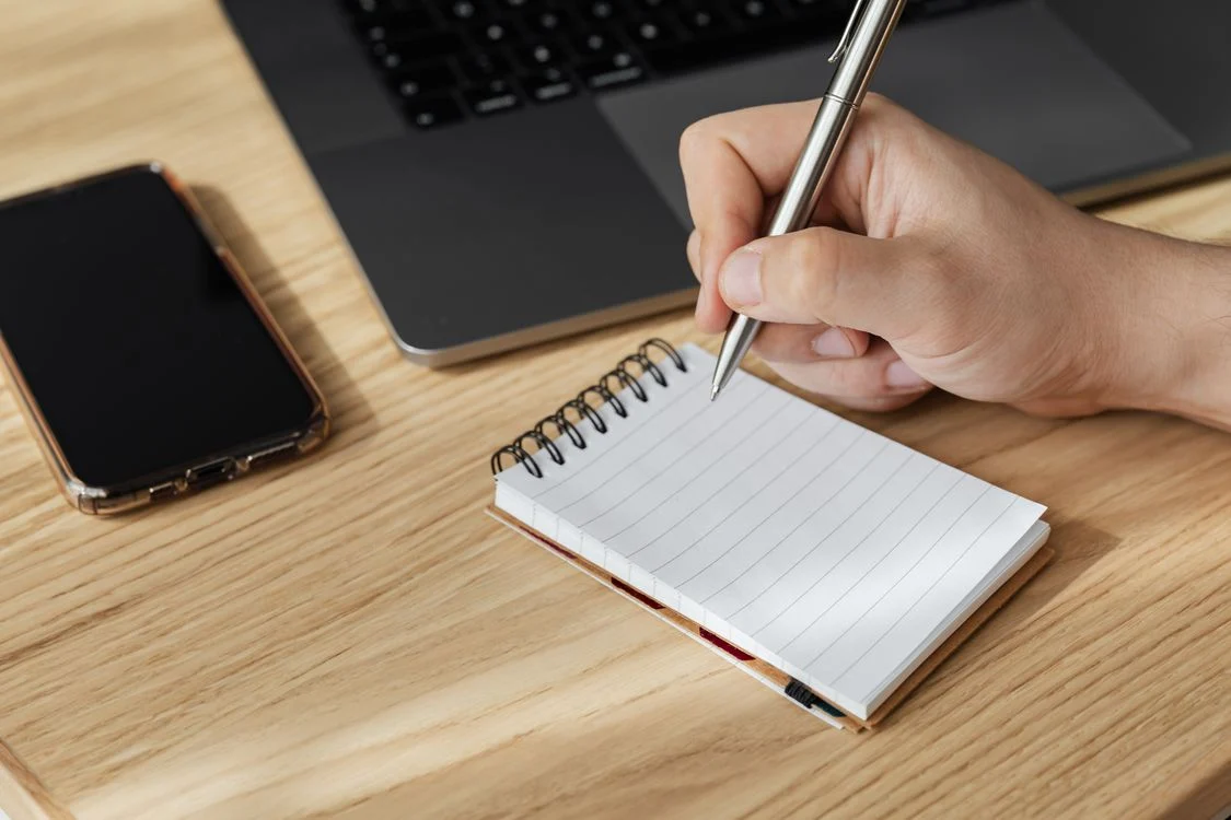 Granola AI Meeting Notes - Hand writing quick notes in a notebook beside a laptop and phone during a work meeting